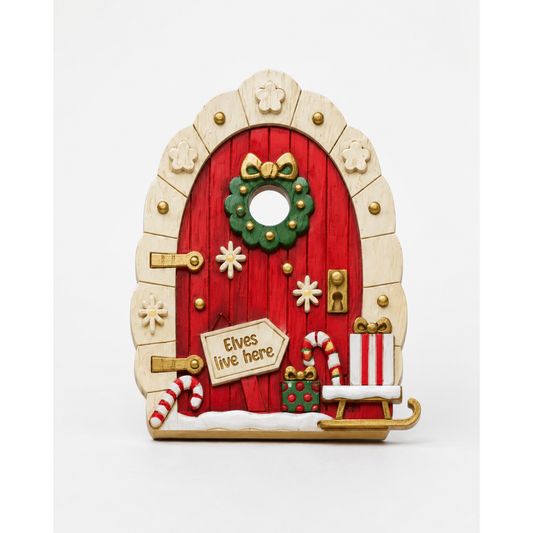 A decorative Christmas fairy door ornament with a red wooden-textured door, beige arched frame, green wreath, gold accents, candy canes, snow, and a sign reading 'Elves live here,' shown on a white background.